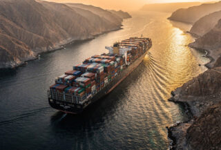 Container ship transiting Strait of Hormuz at dawn, narrow waterway between Iran and Oman