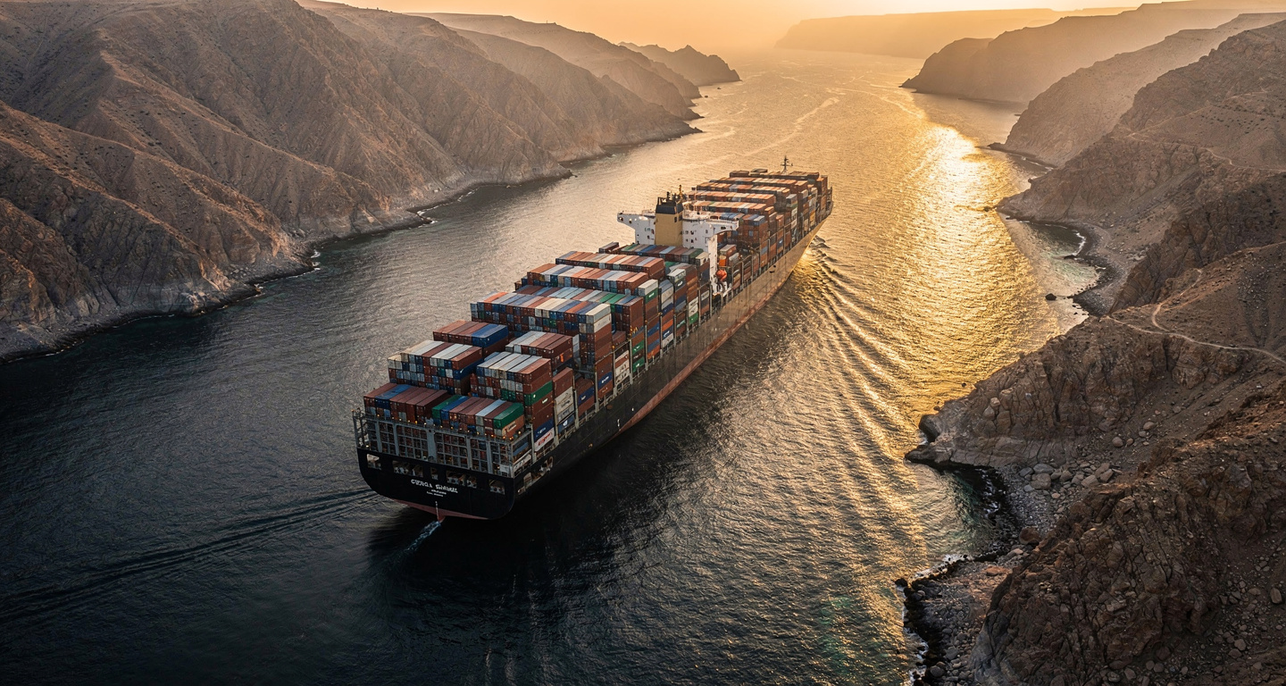 Container ship transiting Strait of Hormuz at dawn, narrow waterway between Iran and Oman