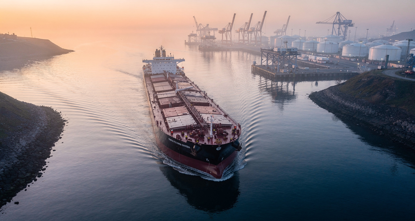 Oil tanker in narrow strait with port infrastructure, dawn lighting, aerial view