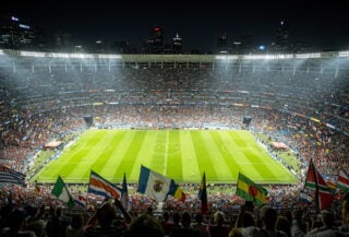 Packed football stadium at night with fans holding Asian national flags under dramatic lighting