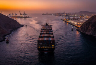 Container ship transiting narrow Red Sea strait with port infrastructure visible