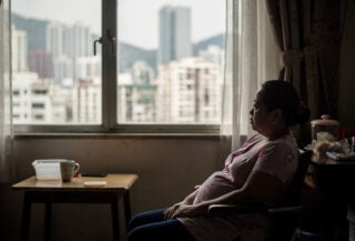Filipino domestic worker in Hong Kong apartment, looking out window at city skyline