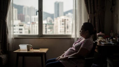 Filipino domestic worker in Hong Kong apartment, looking out window at city skyline