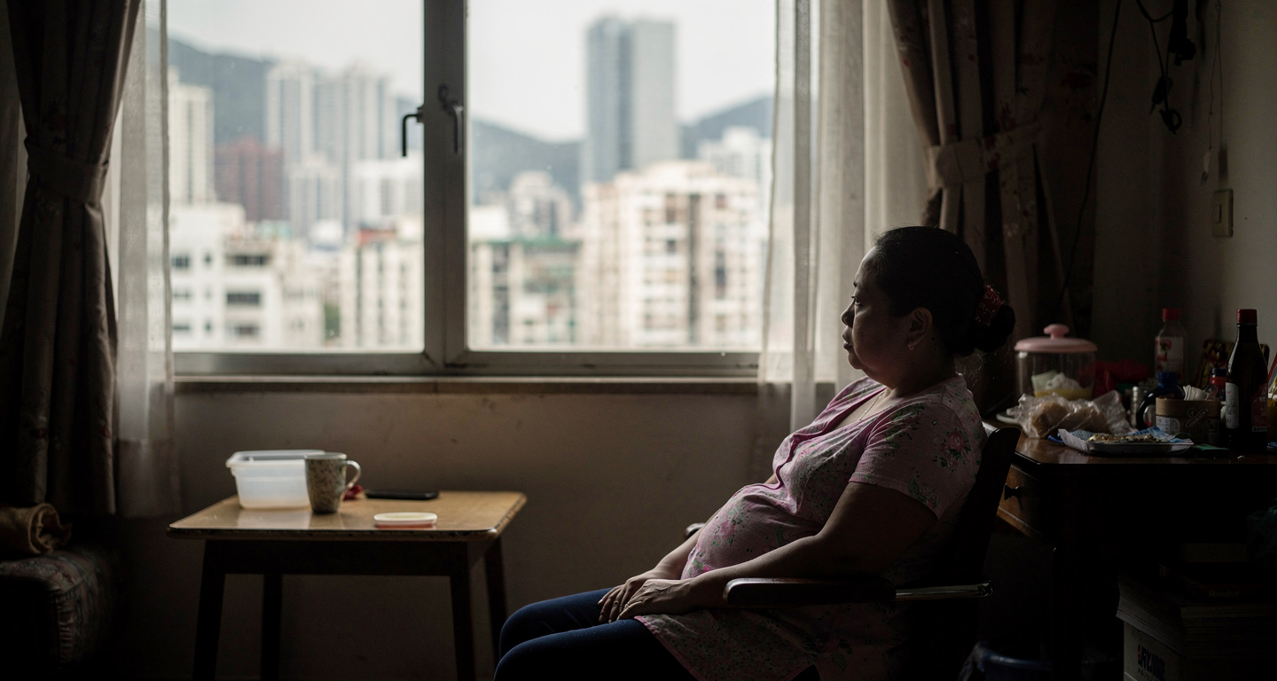 Filipino domestic worker in Hong Kong apartment, looking out window at city skyline