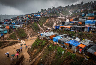 Hillside refugee camp in Cox's Bazar during monsoon rains, steep terrain with makeshift shelters