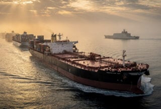 Oil tanker in Strait of Hormuz with military presence and queued container ships at dawn