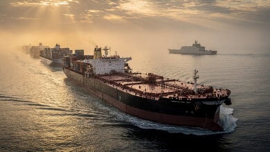 Oil tanker in Strait of Hormuz with military presence and queued container ships at dawn