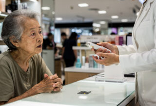 Elderly woman at beauty counter in Hong Kong department store, salesperson demonstrating skincare device, credit card on counter