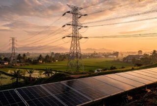 High-voltage transmission towers and power lines crossing Southeast Asian landscape with solar panels below, dusk lighting
