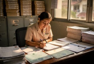 Thai civil registration officer examining birth certificates at Bangkok registration office desk with filing cabinets