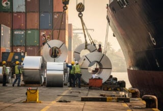 Workers loading steel coils at Jakarta port facility with containers in background, afternoon light