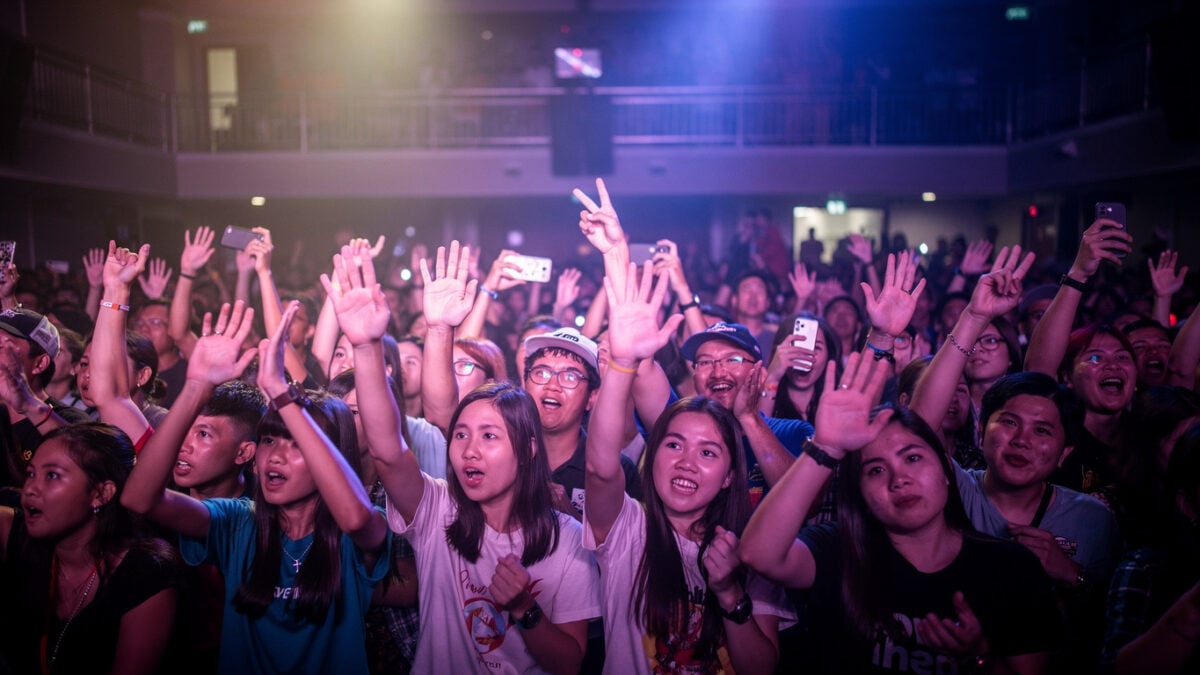 Concert crowd at Bangkok venue with hands raised under colored stage lighting, mixed-age audience