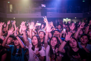 Concert crowd at Bangkok venue with hands raised under colored stage lighting, mixed-age audience