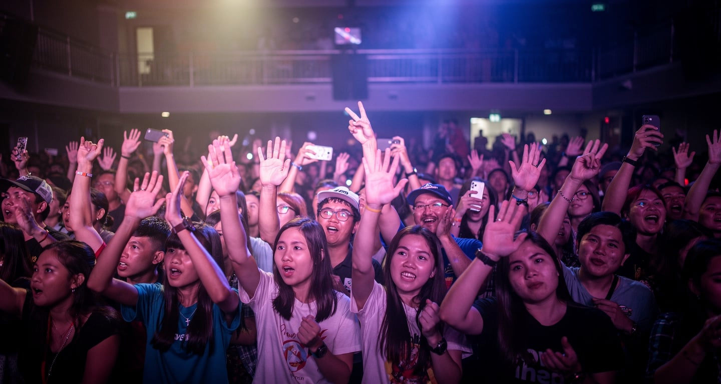 Concert crowd at Bangkok venue with hands raised under colored stage lighting, mixed-age audience