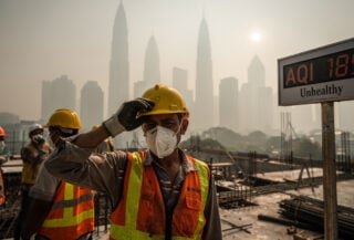 Construction workers in masks on hazy Kuala Lumpur site, one wiping sweat, midday light
