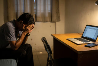 Distressed migrant worker in sparse apartment with laptop, head in hands, representing trafficking victims