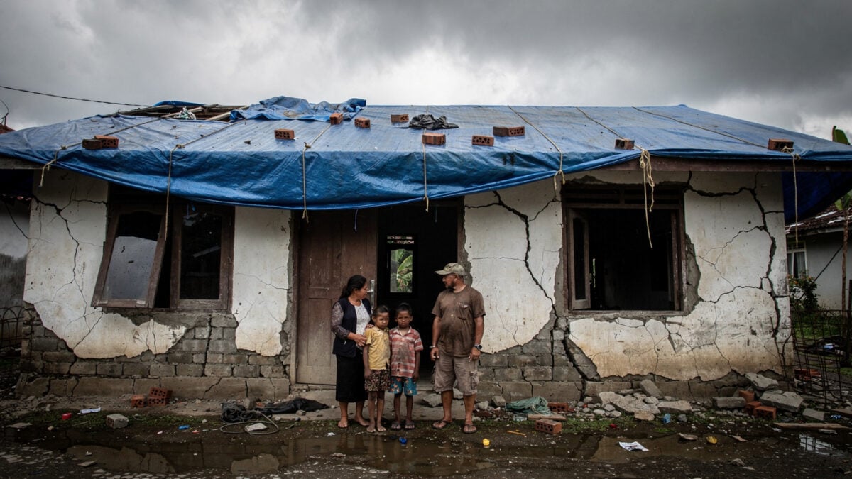 Family outside cracked concrete home in Flores with structural damage and temporary tarp covering, grey sky