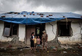 Family outside cracked concrete home in Flores with structural damage and temporary tarp covering, grey sky