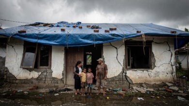 Family outside cracked concrete home in Flores with structural damage and temporary tarp covering, grey sky