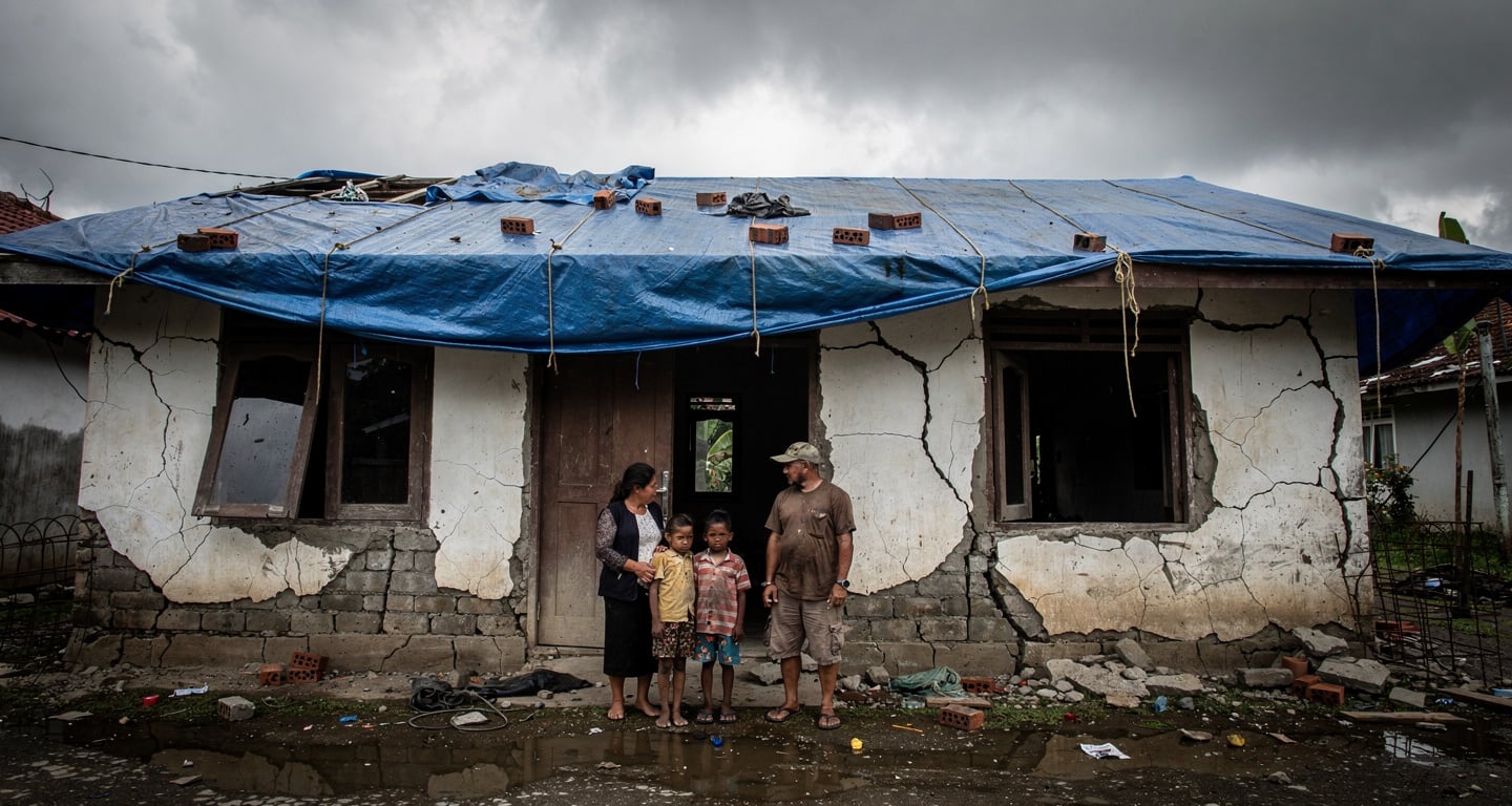 Family outside cracked concrete home in Flores with structural damage and temporary tarp covering, grey sky