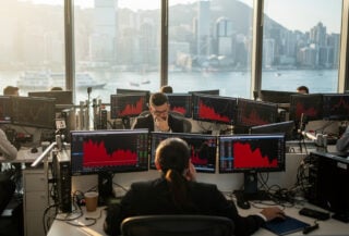 Hong Kong traders monitoring falling stock charts on red screens in dealing room overlooking Victoria Harbour