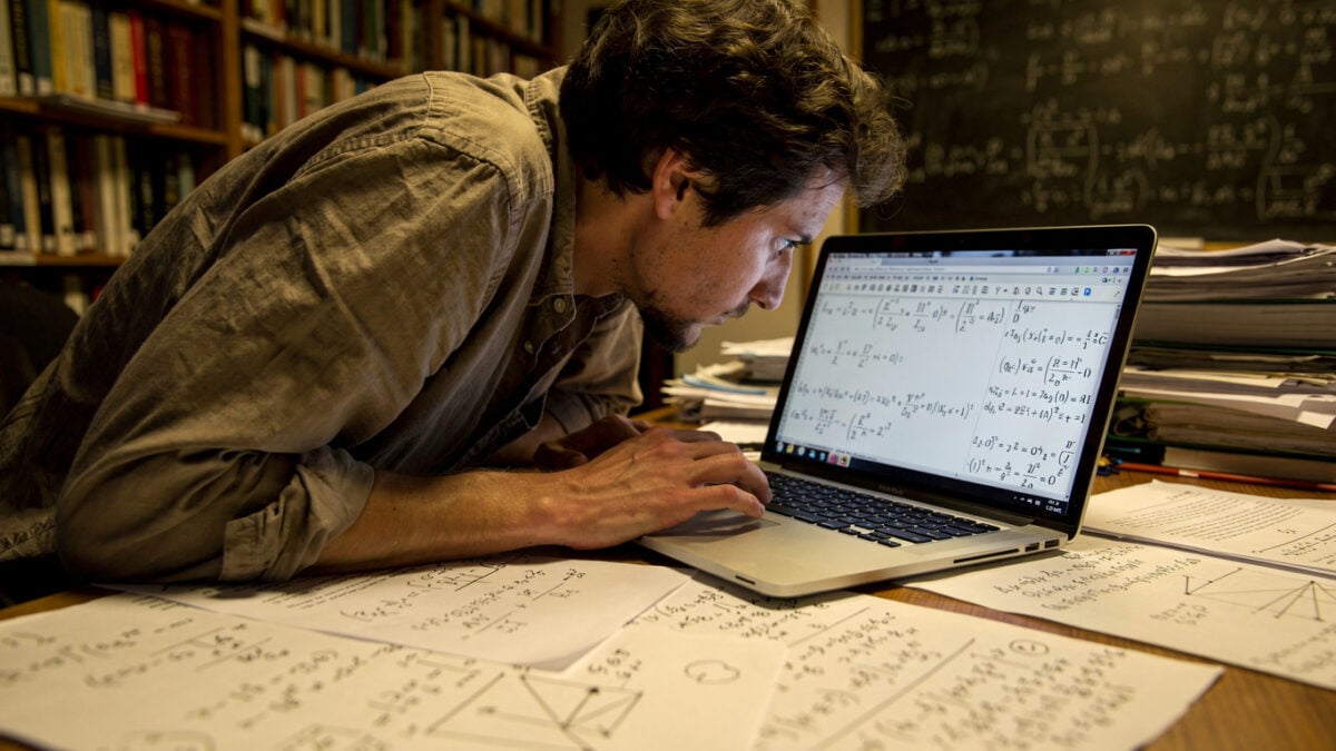 Mathematician studying equations on laptop screen in university office, scattered papers, warm overhead lighting