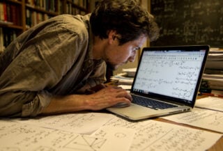 Mathematician studying equations on laptop screen in university office, scattered papers, warm overhead lighting