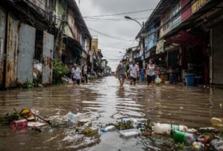 Residents wading through floodwater on Bangkok street during monsoon deluge, shophouses visible