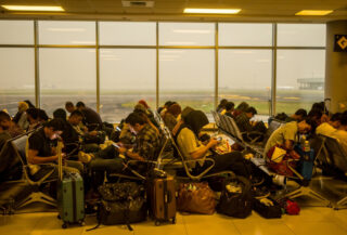 Stranded passengers with luggage in crowded Jakarta airport terminal, volcanic ash haze visible through windows