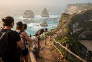 Tourists at Kelingking Beach clifftop viewpoint, steep descent path with wooden railings visible below, Nusa Penida