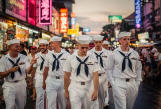 U.S. Navy sailors in dress whites on a crowded Pattaya street at dusk, neon signs glowing
