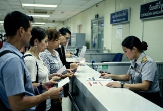 Western tourists reviewing documents at Bangkok immigration counter with Thai official, fluorescent lighting